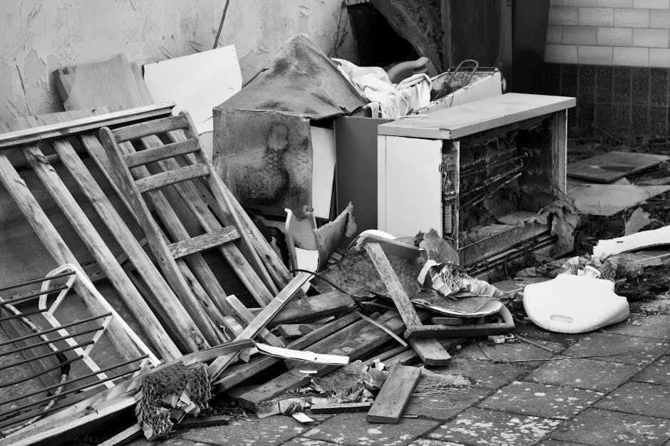 A pile of discarded household items and debris is depicted outdoors on a paved surface, adjacent to a brick wall. The mess includes a weathered wooden garden chair with slatted back and seat, leaning