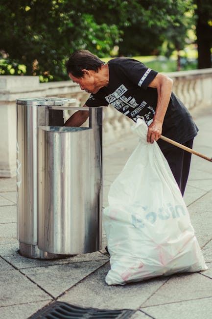 A person's hand holding a green and blue plastic rubbish bag tightly by the tied end, with the bag filled with waste materials, against a plain beige background. The bag is slightly translucent, revea
